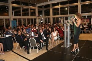 group of people in chairs at tables listening to a woman wearing a green dress at a podium speaking into a mic to the audience
